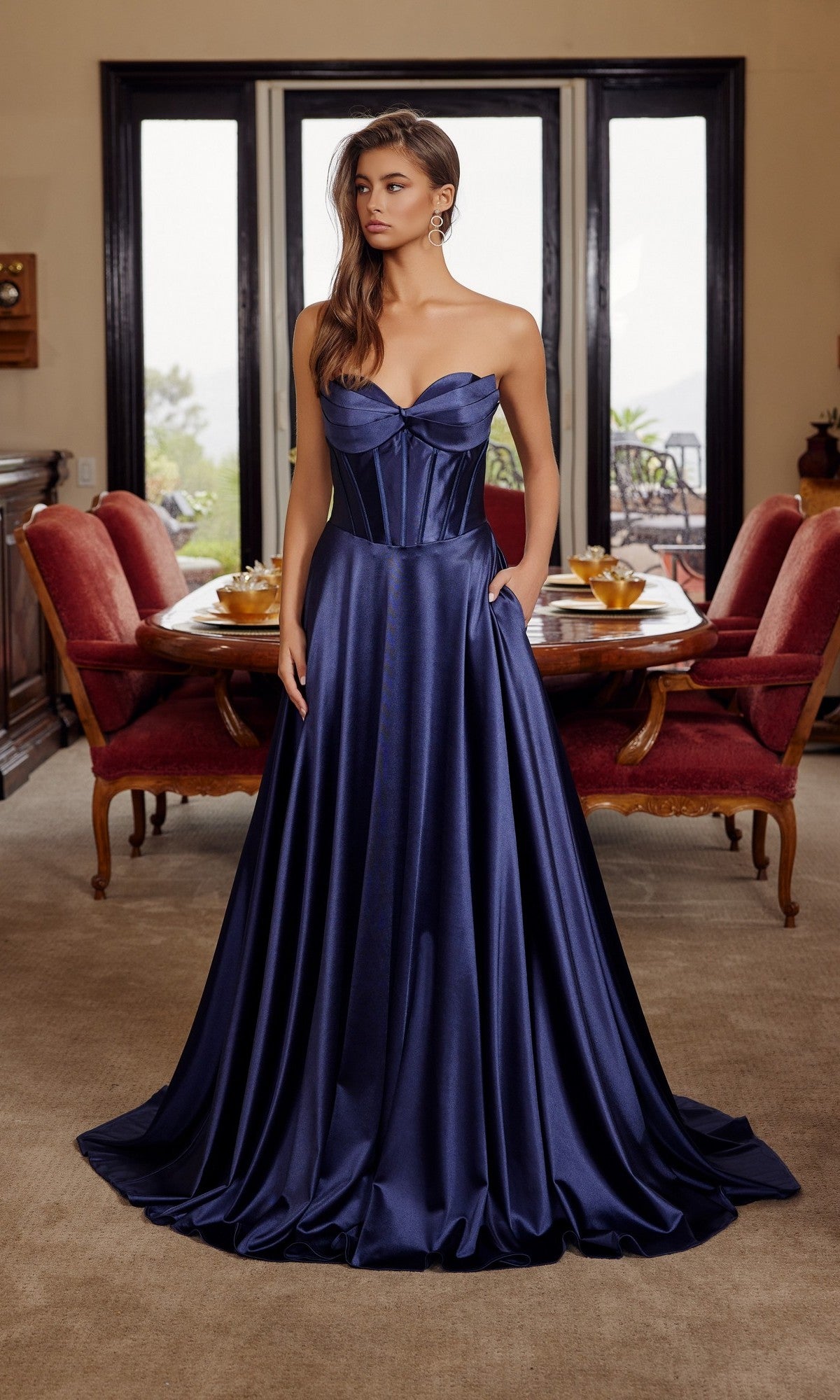 A woman in a navy blue Nox Anabel K1735 long prom dress, featuring a strapless satin corset bodice and elegant A-line skirt, stands in a formal dining room with red chairs and a round table.